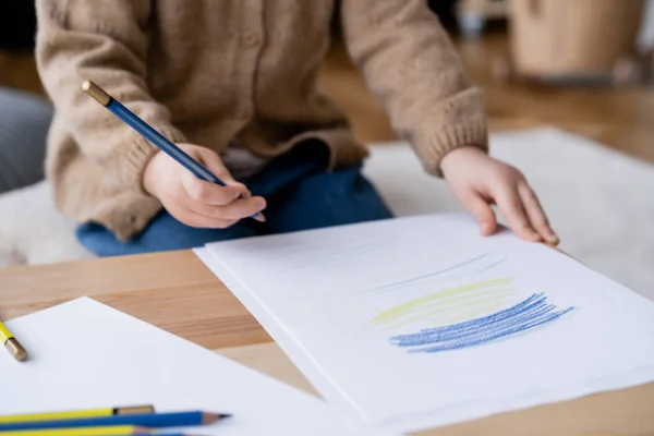 Cropped view of a blurred child holding color pencil near papers with blue and yellow strokes — Stock Photo