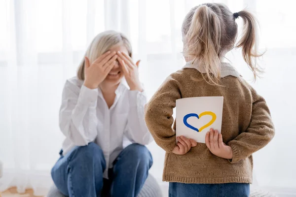 Girl with ponytails holding card with blue and yellow heart near happy mom obscuring face with hands — Stock Photo
