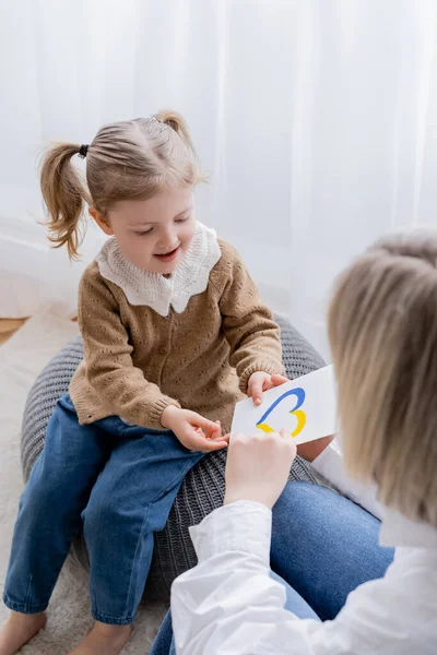 Smiling girl with ponytails holding card with blue and yellow heart near blurred mom — Stock Photo