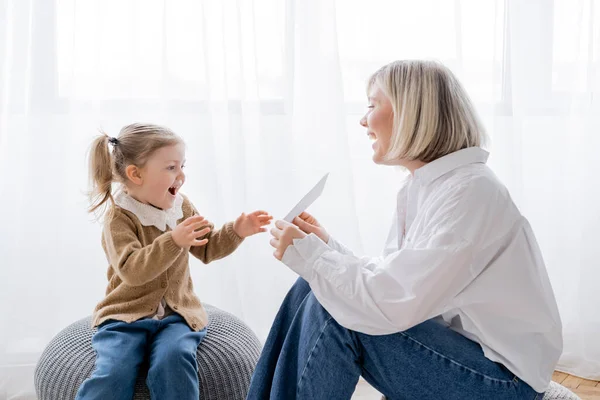 Cheerful blonde woman holding card near amazed daughter while sitting on poufs at home — Stock Photo