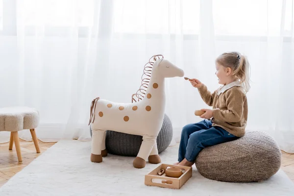 Visão de comprimento total da menina alimentando cavalo de brinquedo de tigela de madeira enquanto joga no pufe em casa — Fotografia de Stock