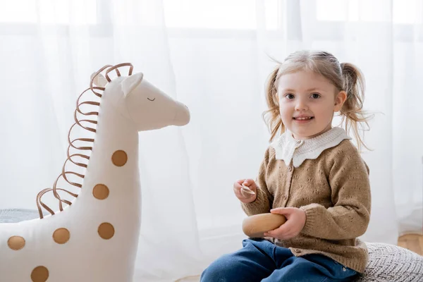 Little girl smiling at camera while holding wooden spoon and bowl near toy horse — Stock Photo