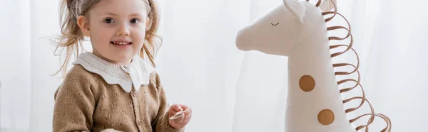 Fille heureuse avec cuillère en bois regardant la caméra près du cheval jouet, bannière — Photo de stock