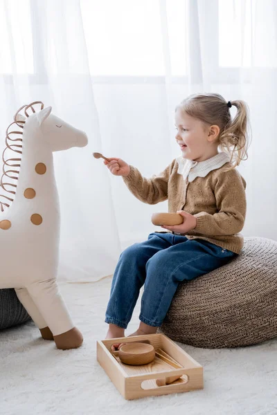 Full length view of little girl sitting on pouf and feeding toy horse from wooden bowl — Stock Photo