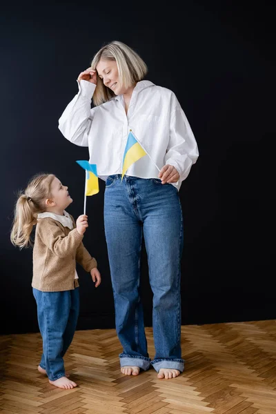 Full length view of barefoot mother and daughter with small ukrainian flags smiling at each other near black wall — Stock Photo