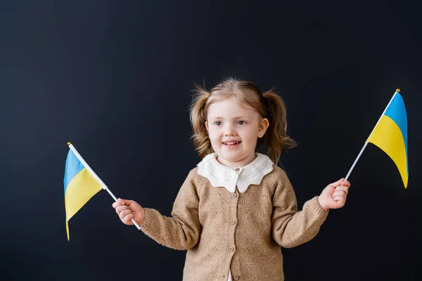 Fröhliches patriotisches Mädchen mit Pferdeschwänzen, die kleine ukrainische Fahnen auf schwarzem Grund schwenken — Stockfoto