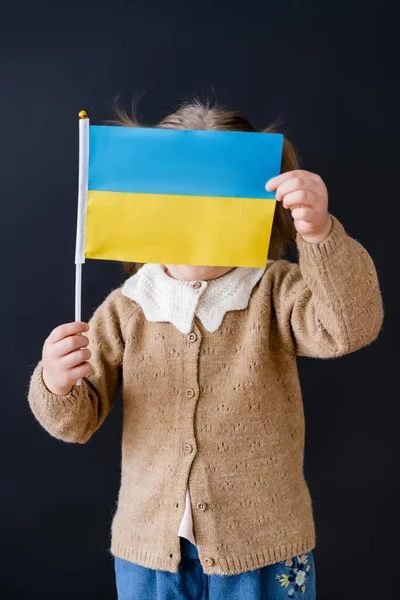 Menina patriótica obscurecendo rosto com pequena bandeira ucraniana isolada no preto — Fotografia de Stock