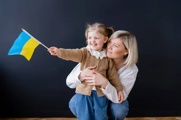 Donna felice abbracciando figlia allegra con bandiera ucraina e guardando lontano vicino al muro nero — Foto stock