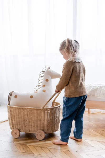 Visão de comprimento total da menina descalça brincando com cavalo de brinquedo no carrinho de vime em casa — Fotografia de Stock