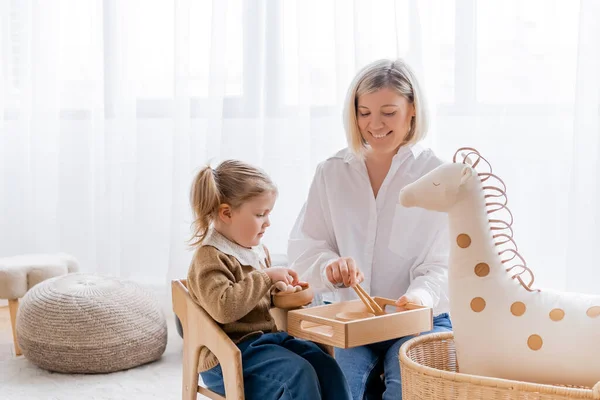 Feliz madre e hija jugando con cuencos de madera y palos cerca de caballo de juguete en casa - foto de stock