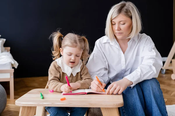 Маленькая девочка с хвостиком рисует с мамой в копировальной книге дома — стоковое фото
