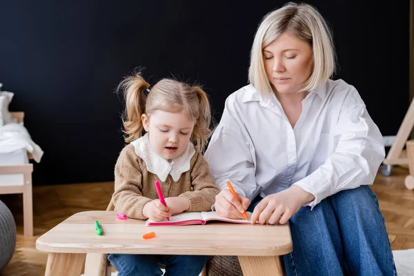 Petite fille avec des queues de cheval dessin avec mère dans le livre de copie à la maison — Photo de stock