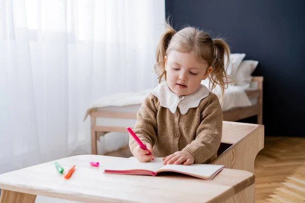 Kleines Mädchen zeichnet mit bunten Filzstiften, während es am Tisch im Schlafzimmer sitzt — Stockfoto