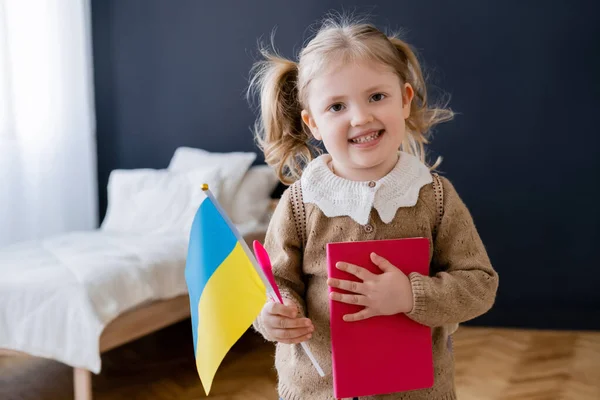 Niño alegre sosteniendo pequeña bandera ucraniana y libro mientras sonríe a la cámara en casa - foto de stock