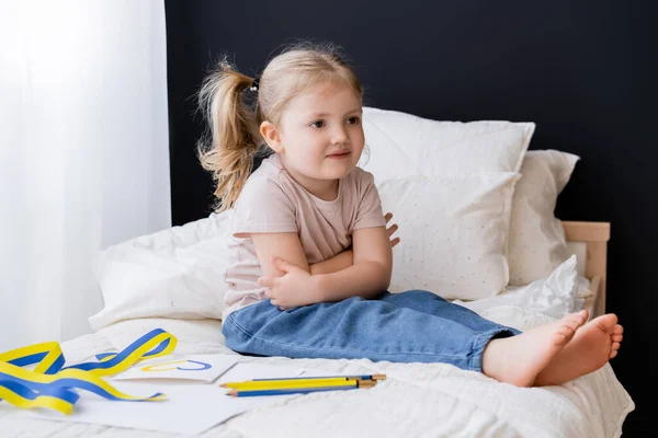 Barefoot girl sitting with crossed arms near blue and yellow ribbon and color pencils on bed — Stock Photo