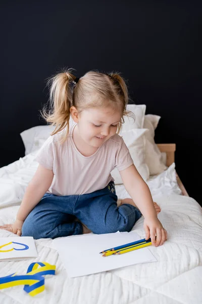 Happy girl sitting on bed near color pencils, ribbon and card with blue and yellow heart — Stock Photo