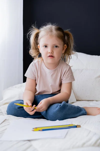 Little girl sitting on bed near blue and yellow pencils and looking at camera — Stock Photo