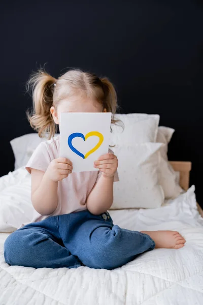 Niña sosteniendo la tarjeta con el corazón azul y amarillo cerca de la cara mientras está sentado en la cama en casa - foto de stock