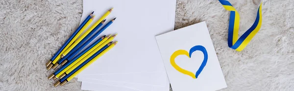 Vue du haut des crayons bleus et jaunes, ruban et carte avec signe du coeur sur tapis moelleux gris, bannière — Photo de stock