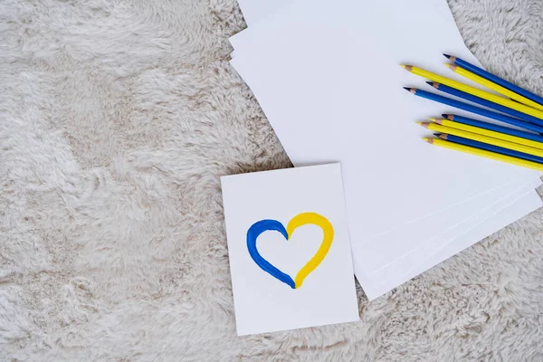 Vue du haut de la carte avec coeur bleu et jaune près des papiers blancs et crayons de couleur sur le tapis — Photo de stock