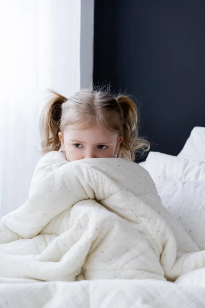 Fille effrayée assis sur le lit et le visage obscurcissant avec couverture blanche — Photo de stock