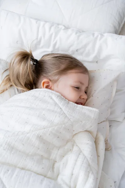 Vista de ángulo alto de niña feliz acostada en la cama bajo una manta blanca - foto de stock