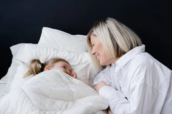 Blondine lächelt neben kleiner Tochter, die unter weißer Decke liegt — Stockfoto