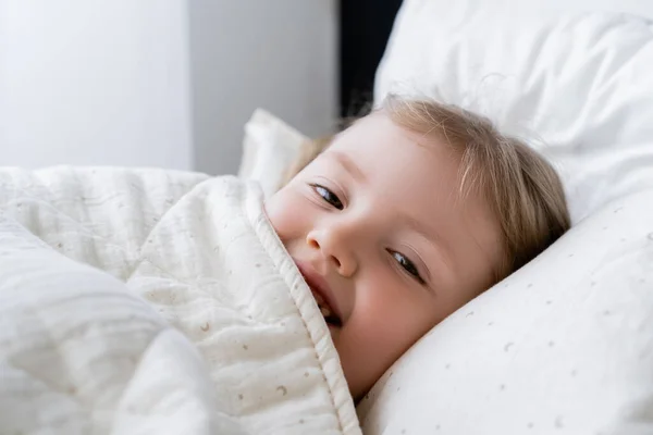 Cheerful child looking at camera while lying in bed under white blanket — Stock Photo