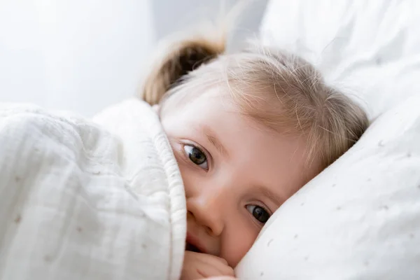 Close up view of little girl lying in bed under blanket and looking at camera — Stock Photo