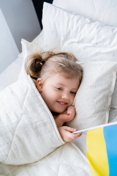 High angle view of smiling patriotic girl lying in bed with small ukrainian flag — Stock Photo