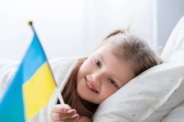 Patriotic girl lying in bed at home and holding small ukrainian flag — Stock Photo