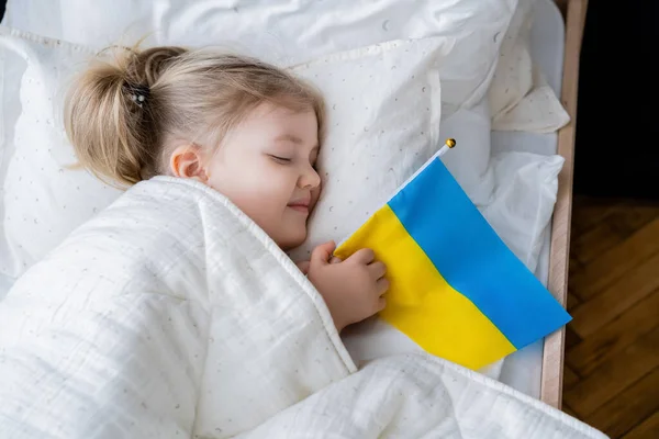 Top view of smiling girl sleeping in bed with small ukrainian flag — Stock Photo