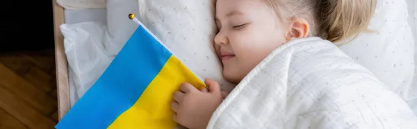 Вид сверху на улыбающуюся девушку, спящую с маленьким украинским флагом в постели, баннер — стоковое фото