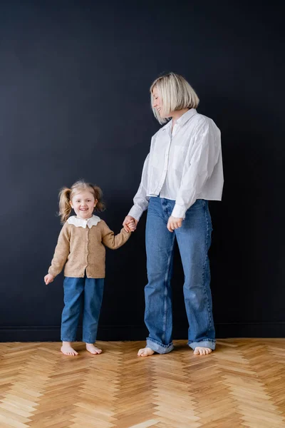 Visão de comprimento total de mãe e filha descalça de mãos dadas perto da parede preta em casa — Fotografia de Stock