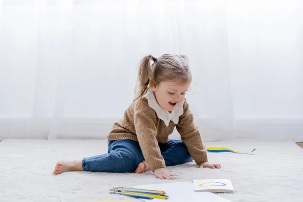 Збуджена дівчина сидить на підлозі біля кольорових олівців і листівки з синьо-жовтим серцем — стокове фото