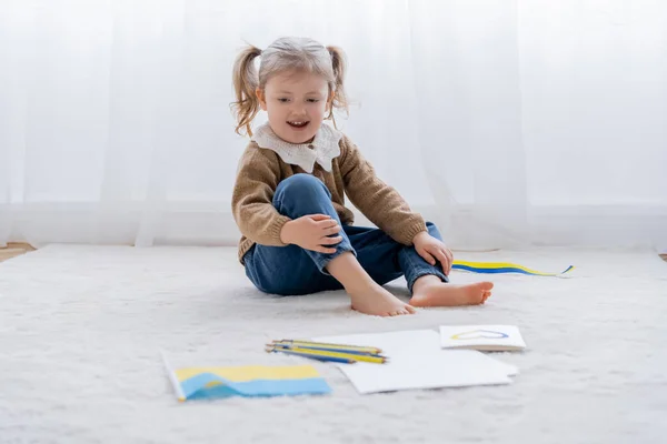 Barefoot girl sitting on floor near ukrainian flag, color pencils and card with blue and yellow heart — Stock Photo