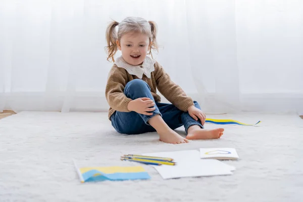 Fille pieds nus assis sur le sol près du drapeau ukrainien, crayons de couleur et carte avec coeur bleu et jaune — Photo de stock