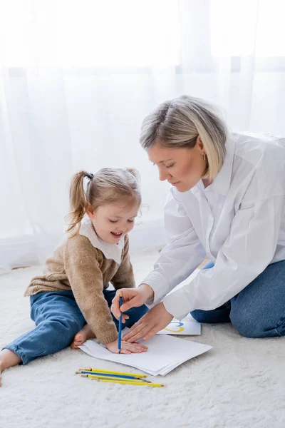 Femme blonde dessin avec crayon bleu près de fille souriante sur le sol à la maison — Photo de stock