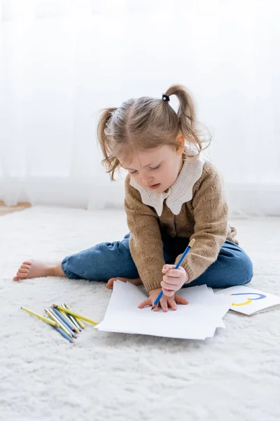 Little girl with ponytails drawing on floor near card with blue and yellow heart — Stock Photo