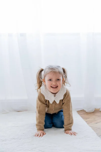 Alegre chica con coletas arrastrándose en el suelo en casa y mirando a la cámara - foto de stock