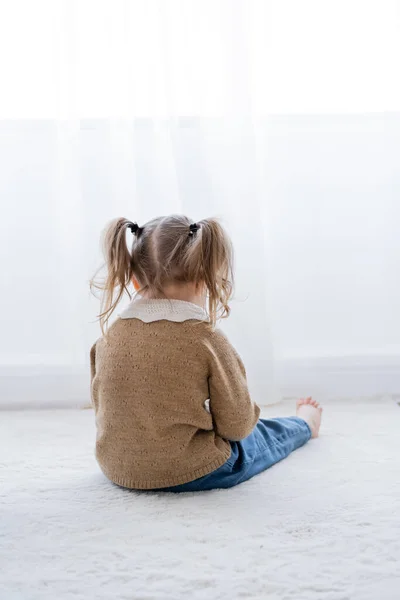 Back view of offended little girl with ponytails sitting on floor at home — Stock Photo