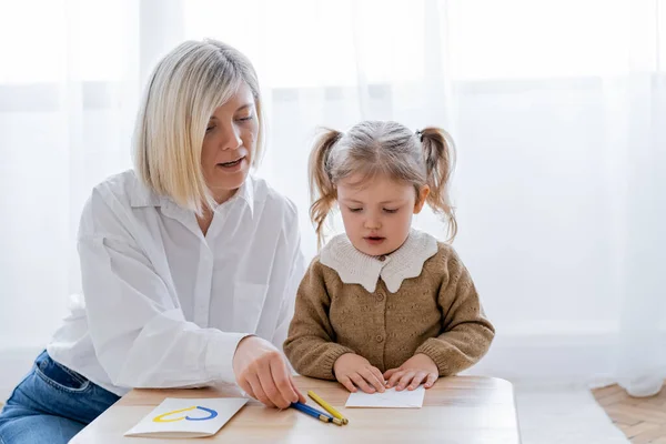 Blonde woman and girl with ponytails sitting near color pencils and card with blue and yellow heart — Stock Photo