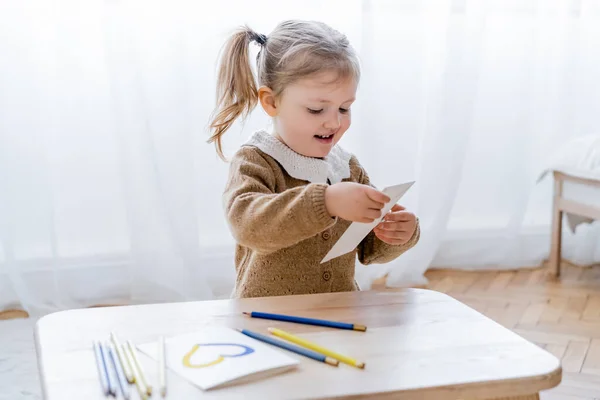 Heureuse fille tenant du papier près de la table avec crayons de couleur et carte avec coeur bleu et jaune — Photo de stock