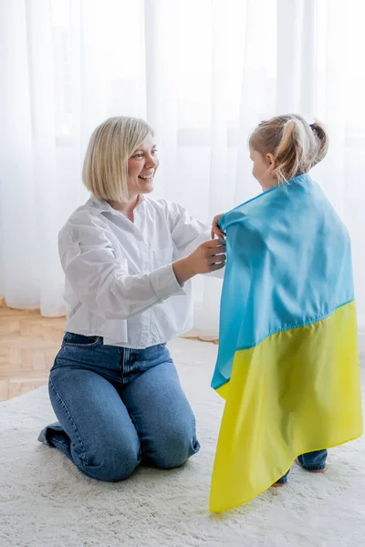 Lächelnde blonde Frau bedeckt kleine Tochter zu Hause mit ukrainischer Flagge — Stockfoto