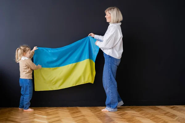 Full length view of patriotic mother and daughter holding ukrainian flag near black wall — Stock Photo
