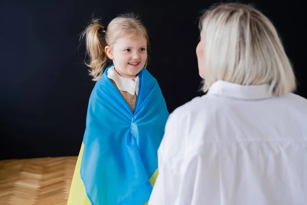 Маленька дівчинка, вкрита прапором українки, посміхається вдома біля розмитої мами. — стокове фото