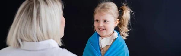 Little girl with ponytail smiling near blurred mother isolated on black, banner — Stock Photo