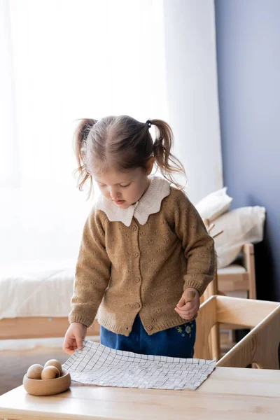 Chica con coletas sosteniendo cuchara de madera y servilleta a cuadros mientras juega en casa - foto de stock