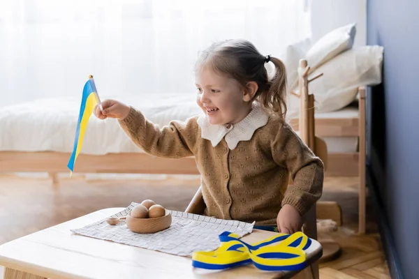 Happy girl holding ukrainian flag near blue and yellow ribbon and wooden toys on desk — Stock Photo