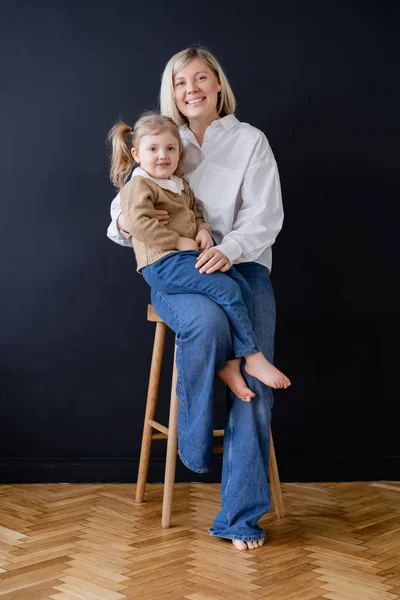 Cheerful woman smiling at camera near daughter sitting on high stool by black wall — Stock Photo